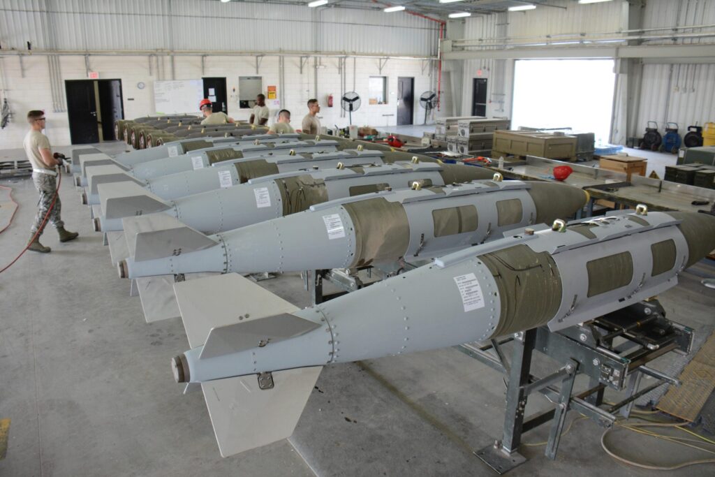 Rows of assembled aerial munitions in a U.S. Air Force maintenance facility, with airmen working on inspection and preparation tasks.